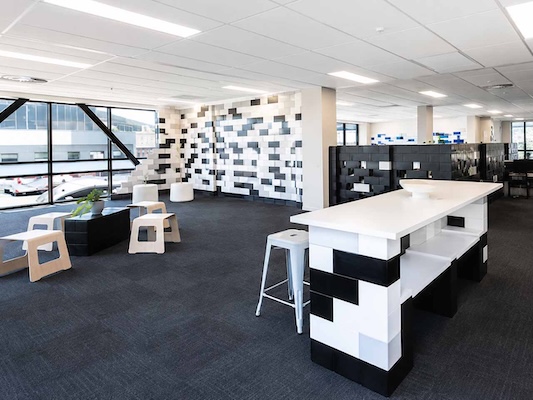 A modern open office with white and black modular desks, stools, seating areas, and large windows, featuring contemporary room dividers and a geometric wall design.