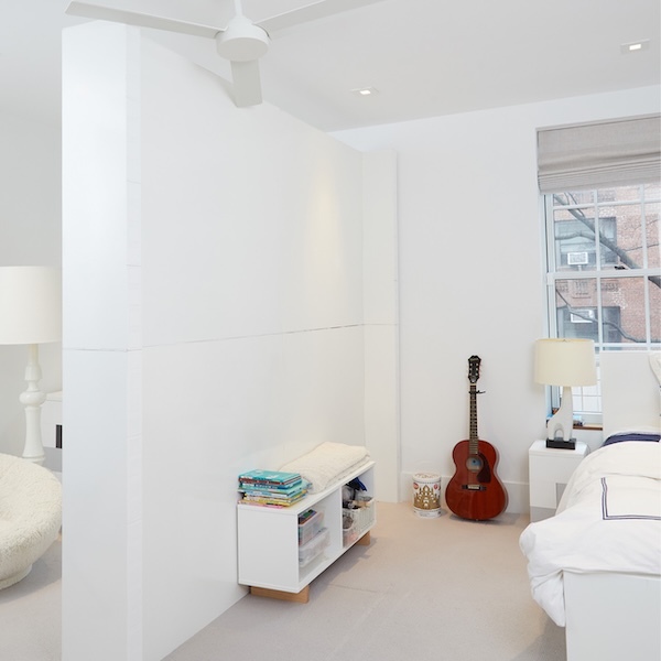 Bright, minimalist bedroom with white walls, a cozy bed, a small storage bench with books, an acoustic guitar in the corner, and natural light streaming through a large window.