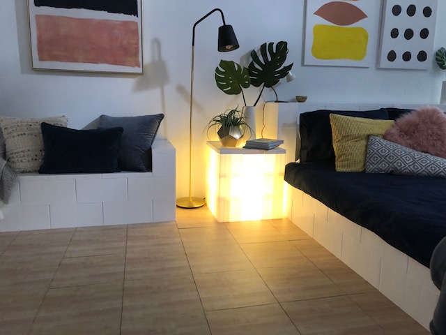 White modular block seating and illuminated side table creating a modern lounge area with plants and decorative pillows.