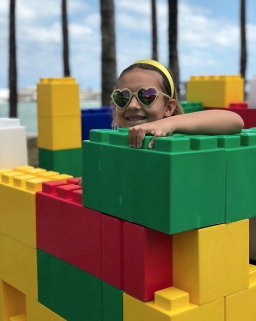 Smiling girl wearing heart-shaped sunglasses peeks over colorful oversized interlocking building blocks.
