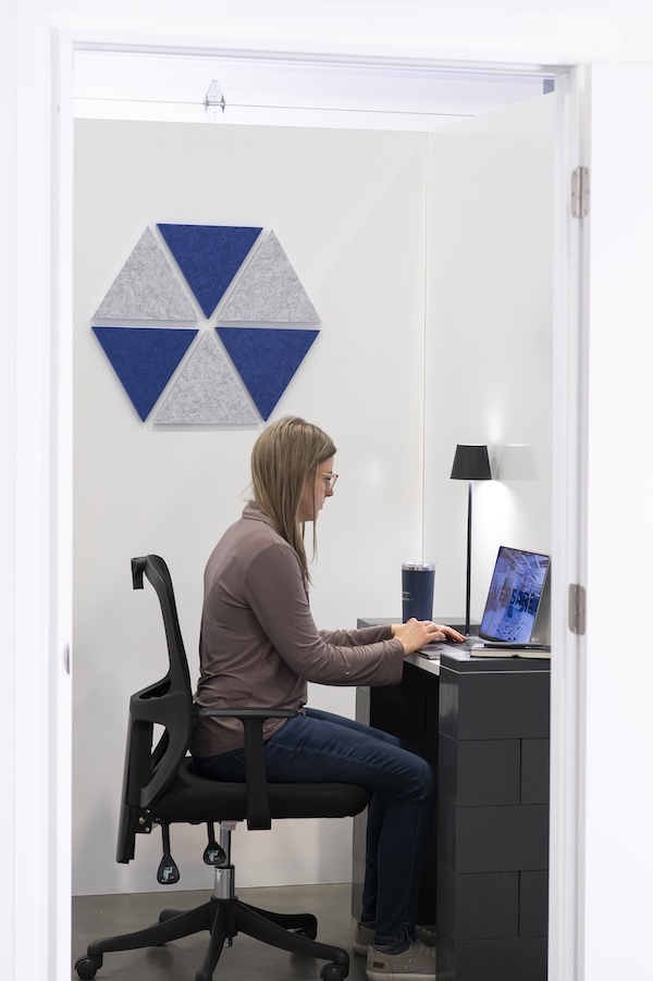 A woman working on a laptop in a compact office booth with blue and gray hexagonal acoustic wall panels behind her.